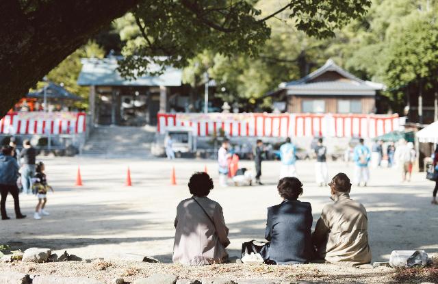 お年寄り3人が座っておまつりを見ている姿を後ろから撮影した写真です