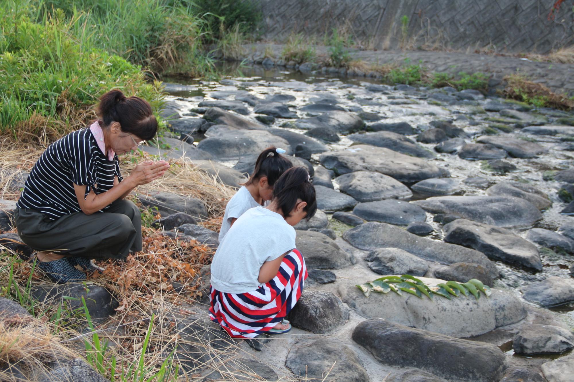 別当川で12枚の柿の葉に炊き込みご飯を乗せたものをお供えし子どもたちが手を合わせている写真