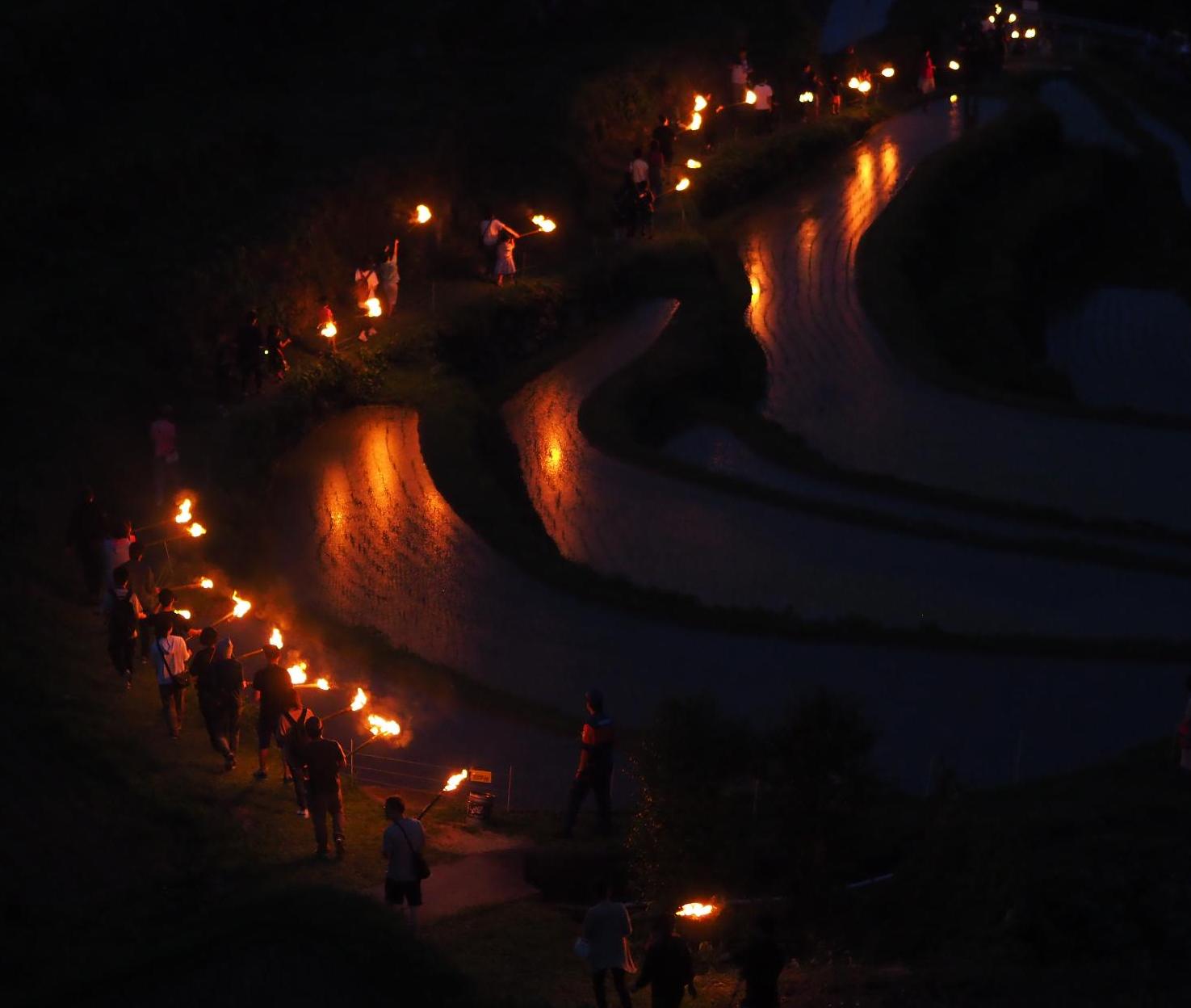 夕暮れの中山千枚田をほてを持った人の列が炎をゆらめかせながら歩いている写真