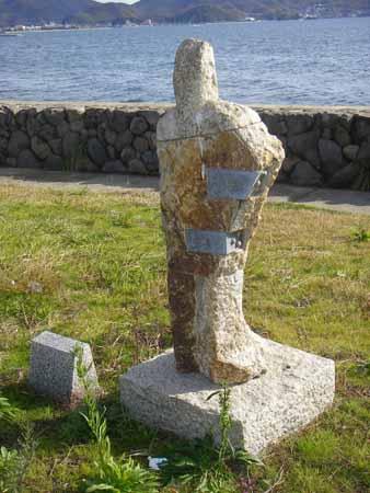 海と石垣で造られた堤防沿いの手前に、四角い石の土台の上に作られた、縦に長く人のような形をした石彫の写真